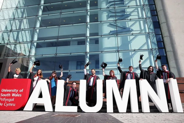 University graduates gathered behind large freestanding ALUMNI letters